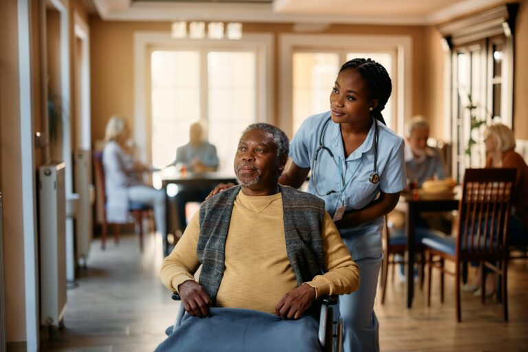 Black nurse and senior man in wheelchair looking through the window at nursing home.